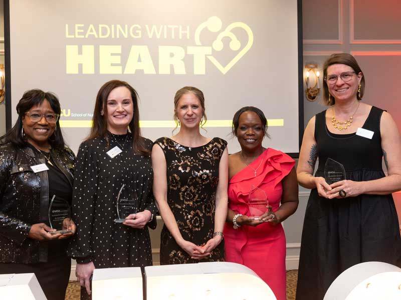 Five award recipients stand side by side holding glass awards at a V.C.U. School of Nursing ceremony, smiling in front of a screen that reads โLeading With Heart.โ