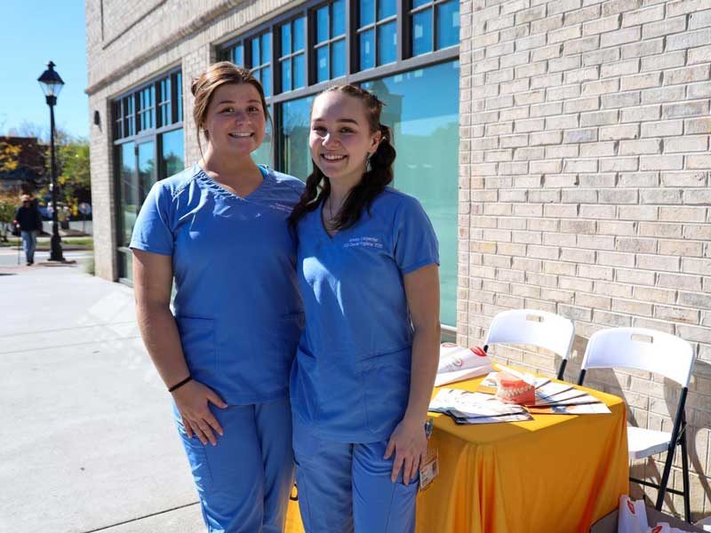 Two students wearing scrubs stand outside a brick building near a table covered with a gold cloth and informational materials