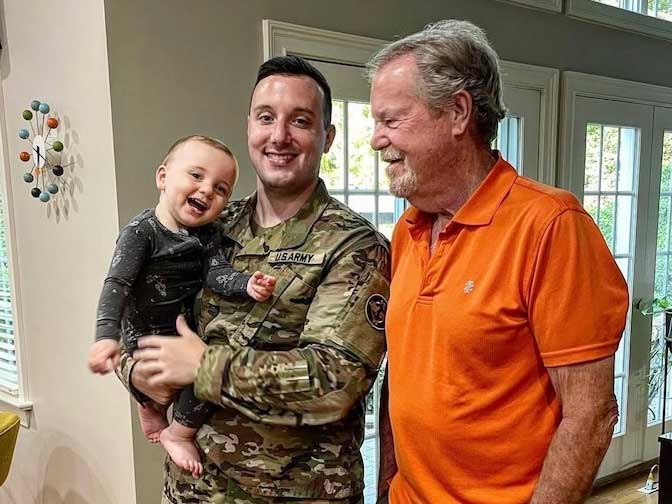 A person in a U.S. Army uniform holds a smiling baby while standing next to an older man inside a home with large windows