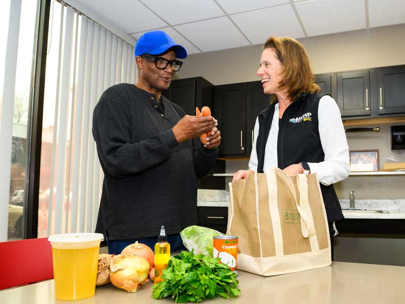 Two adults stand at a kitchen counter talking as one holds several carrots. A grocery bag sits open on the counter beside fresh produce and pantry items.