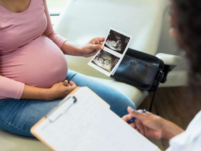 A pregnant woman sitting in a medical office holds ultrasound images of her baby while speaking with a healthcare professional who is taking notes on a clipboard.