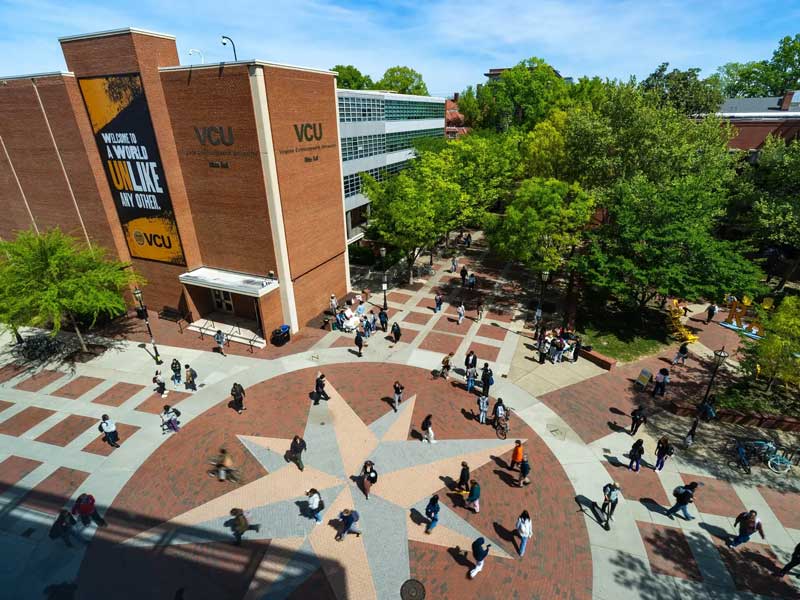 Students walk through a tree-lined V.C.U. campus plaza beside a brick academic building